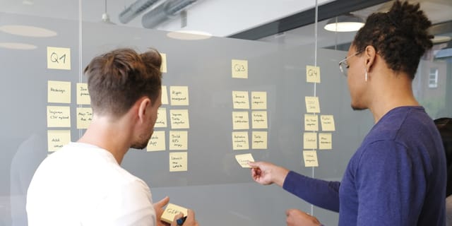 Two people collaborating on a project plan using yellow sticky notes on a glass wall